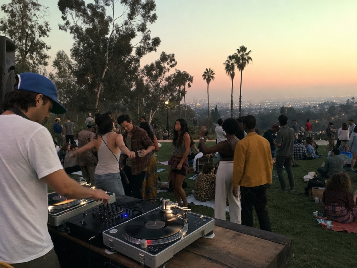 Un DJ tocando vinilos en una reunión en un parque sobre una colina al atardecer, con amigos bailando y sentados en mantas y la ciudad al fondo